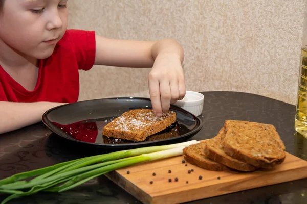 Un niño de 7 años con una camiseta roja prepara un sándwich con poca sal y pan . — Foto de Stock