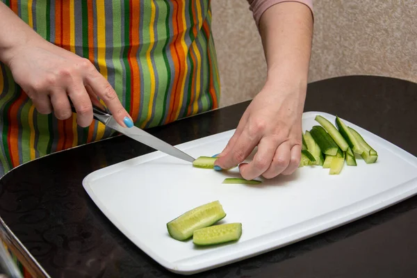 Primer plano manos chica en un delantal a rayas en la cocina está cortando pepinos en tiras en una pizarra blanca en la mesa negra . — Foto de Stock