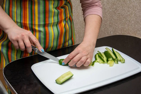 Primer plano manos chica en un delantal a rayas en la cocina está cortando pepinos en tiras en una pizarra blanca en la mesa negra . — Foto de Stock