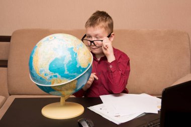 Gözlüklü ve kırmızı gömlekli bir çocuk siyah bir masada oturmuş coğrafi bir küreye bakıyor ve ev ödevi yapıyor. Uzaktan öğrenme konusunda karantinada bir çocuk. Sol gözünde ptosis var..