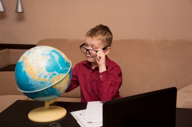 Gözlüklü ve kırmızı gömlekli bir çocuk siyah bir masada oturmuş coğrafi bir küreye bakıyor ve ev ödevi yapıyor. Uzaktan öğrenme konusunda karantinada bir çocuk. Sol gözünde ptosis var..