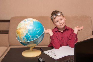 Gözlüklü ve kırmızı gömlekli bir çocuk siyah bir masada oturmuş coğrafi bir küreye bakıyor ve ev ödevi yapıyor. Uzaktan öğrenme konusunda karantinada bir çocuk. Sol gözünde ptosis var..