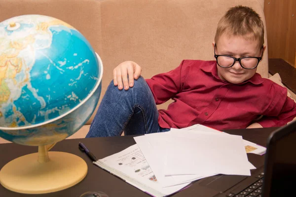 Egy szemüveges fiú és egy piros póló ül egy fekete asztalnál, egy földrajzi földgömböt bámulva és házi feladatot csinálva, egy gyerek karanténban a távoktatásban.. — Stock Fotó