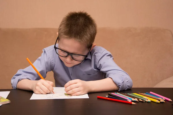 Een 6-7 jarige jongen tekent terwijl hij aan een zwarte tafel zit. Het kind tekent met gekleurde potloden. — Stockfoto