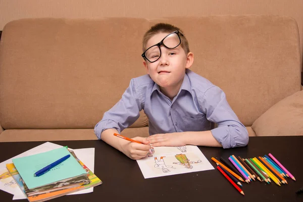 一个带着眼镜的6-7岁男孩坐在一张黑桌边画画。那孩子用彩色铅笔画画. 图库照片