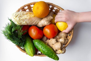 Bir el, içinde portakal, limon, domates, salatalık, dereotu ve zencefil bulunan bir sepetin üzerinde limon tutar..