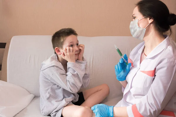 Um médico de casaco branco e luvas médicas azuis segura uma seringa com remédio na frente de um menino, uma criança de pijama cinza tem medo da injeção, chamando um médico em casa. Proteção CONTRA o covid — Fotografia de Stock