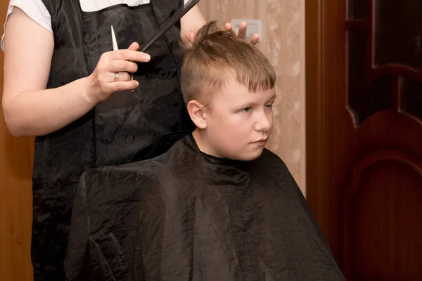 Matka-fryzjer w czarnym fartuchu z grzebieniem i nożyczkami w ręku w domu robi włosy dla syna, który siedzi na krześle i owinięty w czarny koc. Samoizolacja kwarantanny — Zdjęcie stockowe