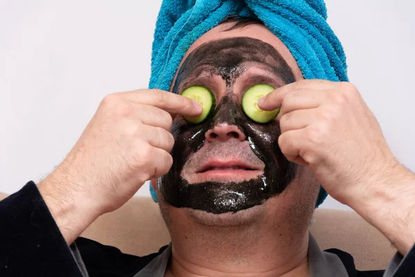 Un concepto divertido de cosmetología. hombre gordo divertido en una bata negra, poniendo rodajas de pepino encima de una máscara de crema negra en su cara, estar de buen humor durante el tratamiento de spa de la mañana . — Foto de Stock