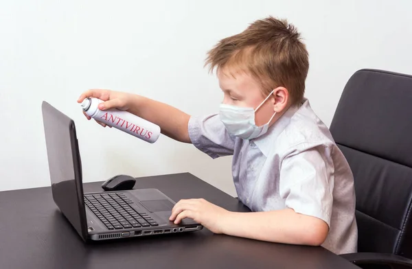 Tıbbi maskeli bir çocuk dizüstü bilgisayarın başında oturuyor ve elinde kırmızı yazıtlı antivirüs olan beyaz bir sprey tutuyor. Siber güvenlik kavramı.