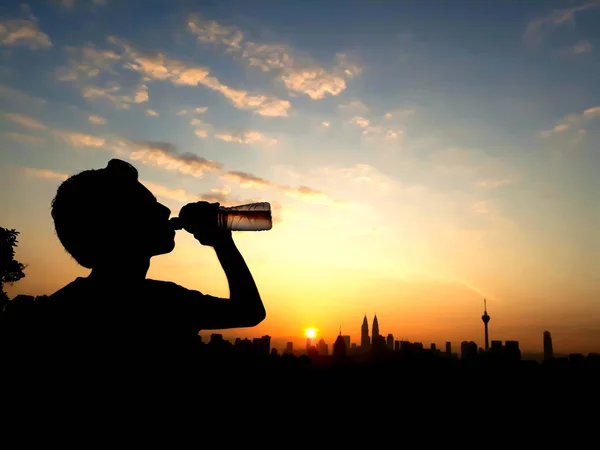 Silhouette shot of young Malay man drinking water during sunrise with building background