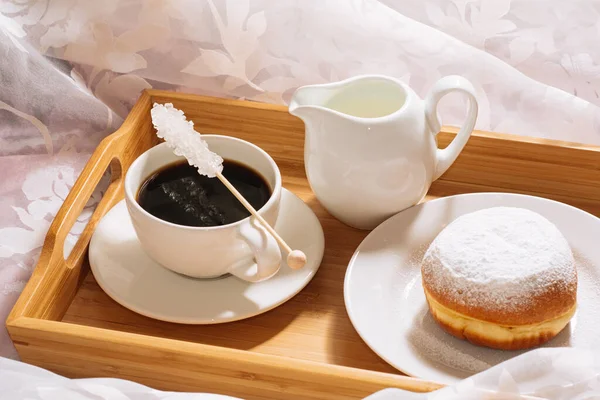 Desayuno por la mañana en la cama con café recién hecho, azúcar cristalina, leche y rosquilla en azúcar en polvo, en una bandeja de madera en platos blancos. De cerca. —  Fotos de Stock