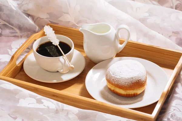 Brunch a letto con caffè fresco tostato e caldo, brocca di latte, ciambella in zucchero a velo su un vassoio di legno in piatti bianchi — Foto Stock