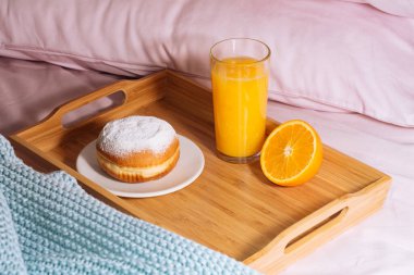 Sabah kahvaltısı taze portakal suyu, yarım portakal ve pudra şekerli çörek ahşap bir tepside pembe yastık ve mavi bir ekose ile yatakta.