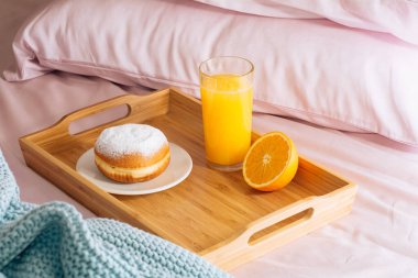 Sabah kahvaltısı taze portakal suyu, yarım portakal ve pudra şekerli çörek ahşap bir tepside pembe yastık ve mavi bir ekose ile yatakta..