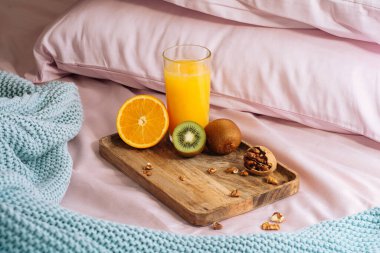 Yatakta sabah sağlıklı kahvaltı taze sıkılmış portakal suyu, yarım portakal, tatlı kivi ve ahşap tepside ceviz dilimleri pembe bir yatak, yastık ve mavi ekose karyola