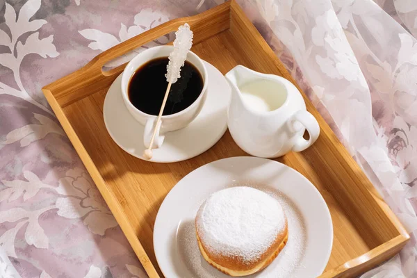 Desayuno en la cama con café caliente y delicioso, leche fresca, azúcar en un palo y una rosquilla de azúcar en polvo en una bandeja de madera en platos blancos —  Fotos de Stock