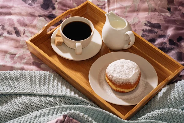 Desayuno en una acogedora cama con café recién hecho, sabroso y fragante, azúcar de caña, leche fresca y rosquilla deliciosa en azúcar en polvo sobre un telón de fondo de sábanas púrpuras y cuadros azules —  Fotos de Stock