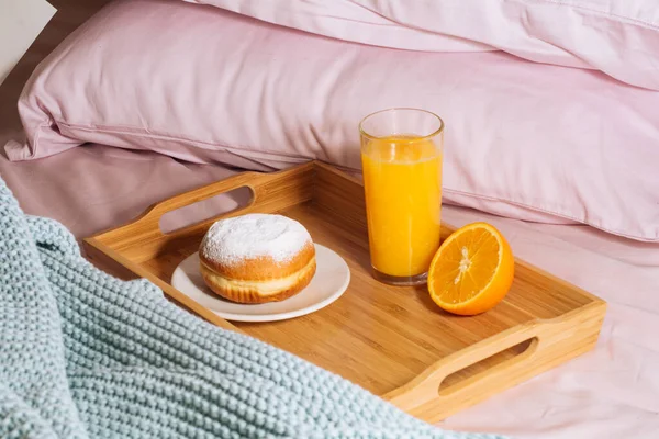 Desayuno por la mañana en la cama con jugo de naranja fresco y saludable y vitamina, media naranja y donut en polvo de azúcar en una bandeja de madera sobre un telón de fondo de almohadas de color rosa y un cuadros azules —  Fotos de Stock