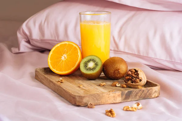Un desayuno saludable en la cama con zumo de naranja fresco, jugosas rebanadas de naranja, kiwi dulce y nuez en una bandeja de madera con un fondo de ropa de cama rosa y almohadas — Foto de Stock