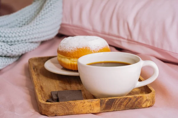 Πρωινό πρωί στο κρεβάτι με φρέσκο και ζεστό καφέ, ένα ντόνατ σε σκόνη ζάχαρης και τρεις φέτες σοκολάτας σε ένα ξύλινο δίσκο με φόντο ροζ σεντόνια και μπλε καρό — Φωτογραφία Αρχείου