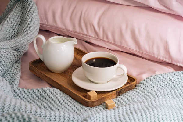 Πρωινό στο κρεβάτι με φρέσκο, αρωματικό και νόστιμο καφέ, μια κανάτα κρύο γάλα και ζάχαρη από ζαχαροκάλαμο σε ένα ξύλινο δίσκο με φόντο ροζ σεντόνια, μαξιλάρια και ένα μπλε καρό — Φωτογραφία Αρχείου