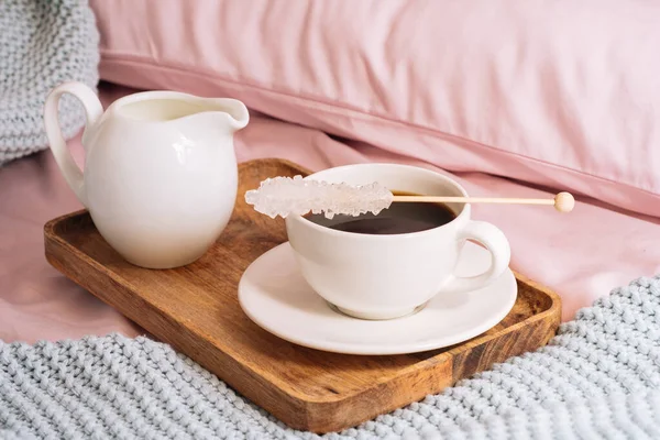 Πρωινό στο κρεβάτι με φρέσκο και νόστιμο καφέ, μια κανάτα κρύο γάλα και κρυσταλλική ζάχαρη σε ένα ραβδί σε ένα ξύλινο δίσκο με φόντο ροζ σεντόνια, μαξιλάρια και ένα μπλε καρό — Φωτογραφία Αρχείου
