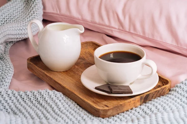 Πρωινό στο κρεβάτι με φρεσκοψημένο και νόστιμο καφέ, μια κανάτα κρύο γάλα και τρεις φέτες πραγματικής μαύρης σοκολάτας σε ένα ξύλινο δίσκο με φόντο ροζ σεντόνια, μαξιλάρια και ένα μπλε καρό — Φωτογραφία Αρχείου