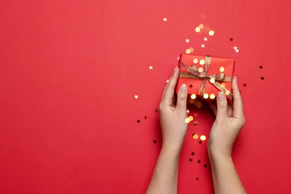 Composición creativa de manos femeninas sosteniendo una caja de regalo roja con arco y brillo estrella caramelo sobre un fondo rojo con lugar para el texto — Foto de Stock