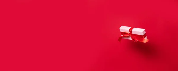 Gyönyörű meglepetés doboz csomagolva rózsaszín papír és egy piros szatén szalag íj szalag szárnyal a levegőben, szemben a piros háttér. Boldog Karácsonyt! Valentin nap. Minimális stílus. — Stock Fotó