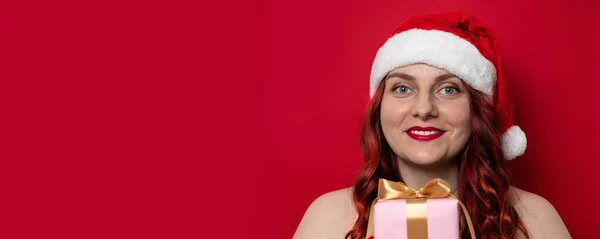 Bella giovane donna con cappello rosso di Babbo Natale con pompon esprimere emozioni felici e tenere presente scatola su sfondo muro rosso — Foto Stock