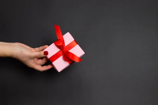 Boldog Ünnepeket koncepció női kéz tartsa meglepetés ajándék piros atlasz szalag sötét háttérrel. Lapos fekvésű, felülnézet — Stock Fotó