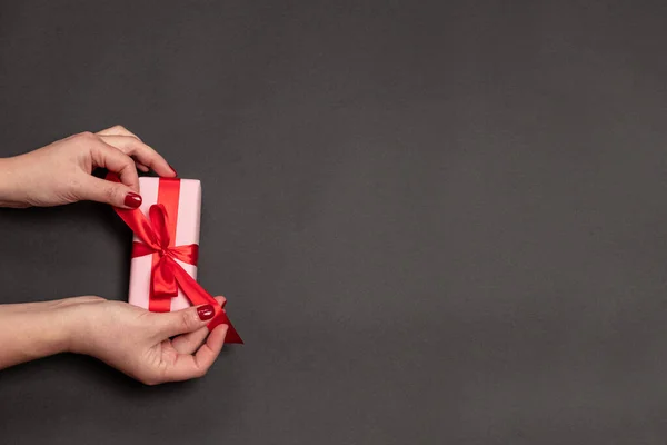 Happy Holidays concept with woman hands holding present box with red bow on pastel dark background . Flat lay style.