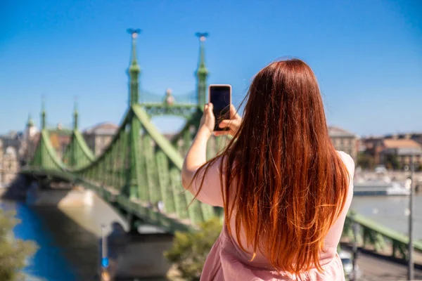 Pembe elbiseli genç kızıl saçlı kız güneşli bir günde köprünün fotoğrafını çeker. — Stok fotoğraf