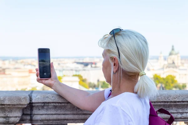 白いTシャツを着た大人の観光客は、旧市街の晴れた日にスマートフォンで街の写真を撮ります. — ストック写真
