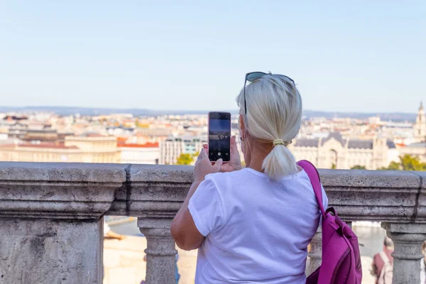 白いTシャツを着た大人のブロンドの女性観光客は、旧市街の晴れた日にスマートフォンで街の写真を撮ります. — ストック写真