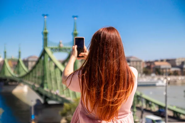 Yazlık pamuk elbiseli güzel kızıl saçlı kız güneşli bir yaz gününde tarihi köprünün fotoğraflarını çekiyor. — Stok fotoğraf