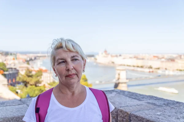 Vuxen blond turist kvinna i en vit T-shirt, med en crimson färg ryggsäck titta på kameran mot bakgrund av den gamla staden Budapest — Stockfoto