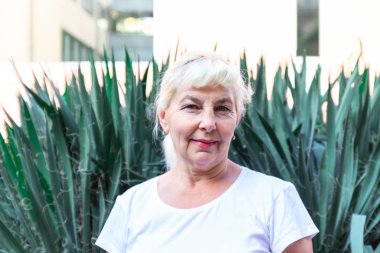 Beyaz tişörtlü yetişkin sarışın kadın yeşil çalıların arkasındaki kameraya bakıp gülümsüyor.