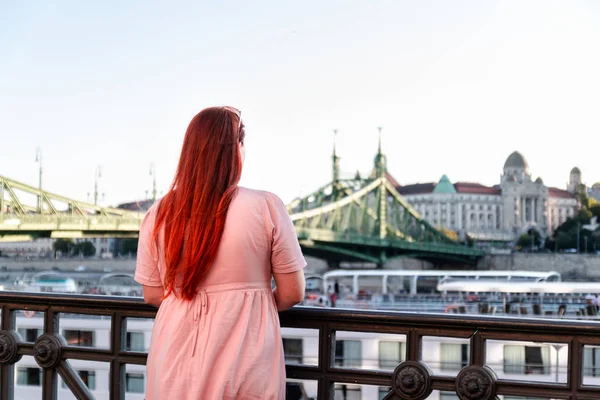 ピンク色の柔らかいドレスを着た赤い髪の少女が川のそばに立って、夕日の鉄の橋を見ています. — ストック写真