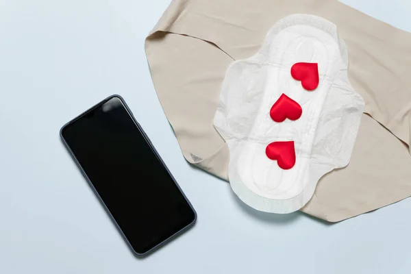 Menstruation Periode Konzept. schlanke Baumwollmenstruationspolster mit roter Liebesform als Blutstropfen, Smartphone auf blauem Hintergrund. — Stockfoto