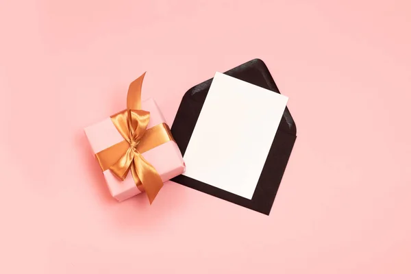 Composición festiva vista superior con hermoso regalo envuelto en papel de vacaciones, cinta de oro y sobre negro con plantilla de papel sobre fondo rosa . — Foto de Stock