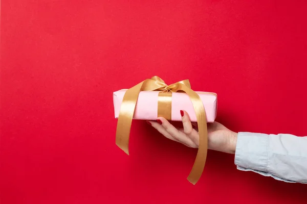 La mano femenina sostiene una envoltura de regalo envuelta en papel rosa y un lazo de cinta de oro sobre un fondo rojo — Foto de Stock