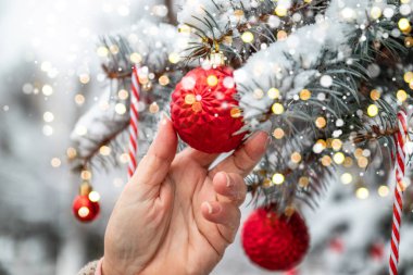 Noel arka planında köknar ağacı dalları, süslemeler ve bulanık ışıklar var. Kadın eli Noel ağacına kırmızı bir top asar.