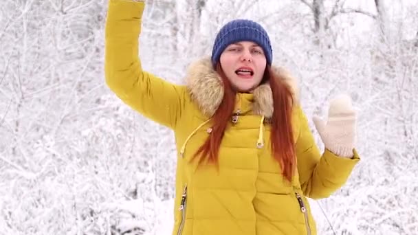 Een jong meisje in een warm geel jasje en een gebreide blauwe hoed en knipt dansend op de achtergrond van een winterlandschap. — Stockvideo