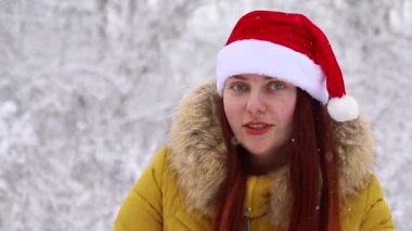Sıcak kış kıyafetleri içinde ve Noel Baba şapkalı genç bir kız ormanda dans ediyor ve bir kış manzarasının arka planında kameraya bakıyor.