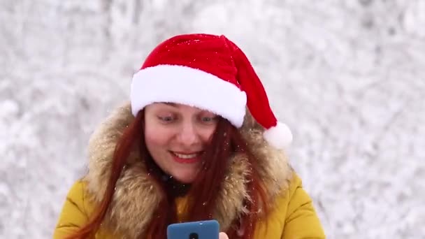 Молодая девушка в теплой зимней одежде и шляпе Санты использует смартфон и испытывает радостные эмоции — стоковое видео