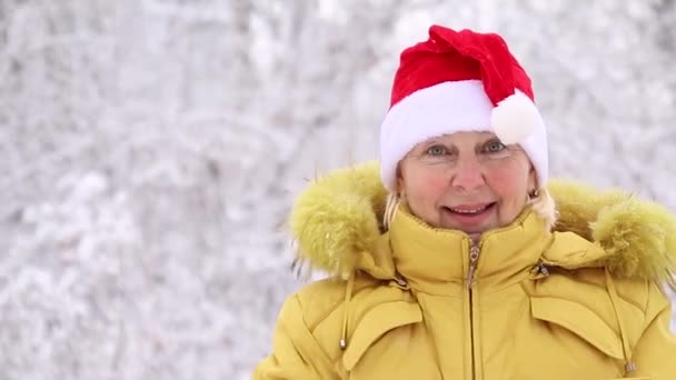 Ενηλίκων γυναίκα με ζεστά φωτεινά ρούχα και καπέλο Σάντα χαμογελά και κοιτάζει την κάμερα — Αρχείο Βίντεο