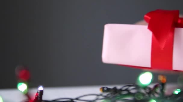 Рука положить упакованный подарочный коробок на стол со светящимися гирляндами на сером фоне — стоковое видео