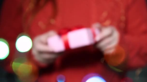 Female hands are holding a surprise gift box with a gold bow and gives a gift. Girl in a red knitted sweater and on background of blurred glitter garlands — Stock Video